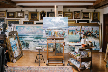 Interior of creative art workshop with painting on easel near shelf with tools and variety of ready framed pictures
