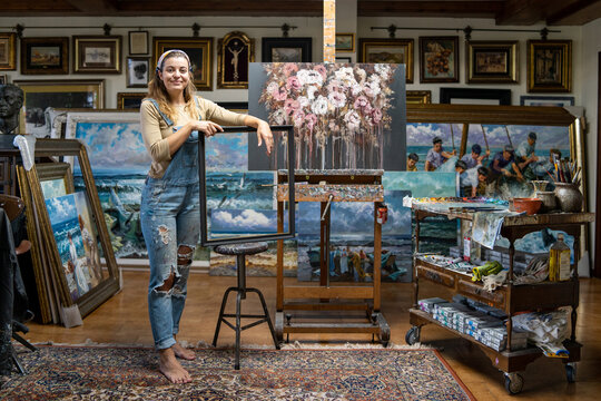 Full length of creative young female artist in denim overall holding empty frame while standing near easel with picture in workshop with paintings