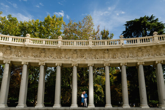 Remote view of couple embracing each other standing on stone columns of arched passage in urban park during summer stroll