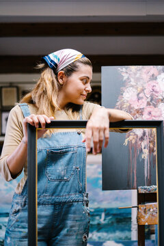 Creative young female artist in denim overall leaning on empty frame in workshop with paintings