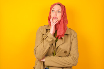 Young caucasian Muslim woman wearing hijab standing against yellow wall covering mouth with hands scared from something or someone bitting nails