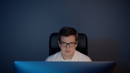 Man in eyeglasses in front of the computer in home office on background of grey wall. Pan shot of young man sitting at home working alone at night