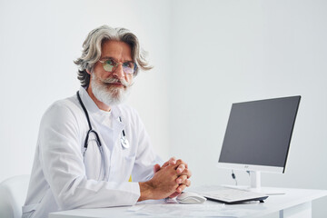 Portrait of senior male doctor with grey hair and beard in white coat is indoors in clinic