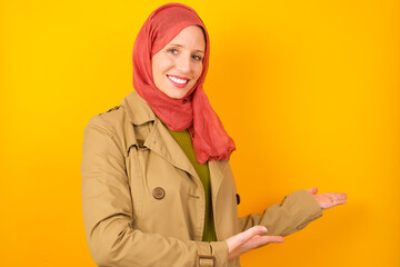 Young caucasian Muslim woman wearing hijab standing against yellow wall Inviting to enter smiling natural with open hands. Welcome sign.