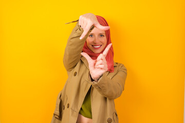 Positive Young caucasian Muslim woman wearing hijab standing against yellow wall with cheerful expression, has good mood, gestures finger frame actively at camera.