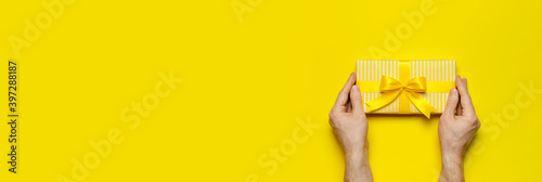 Male hands holding bright yellow gift present box with ribbon on yellow background top view. Flat lay holiday background. Birthday present, March 8, Mother's Day, Valentine's Day. Congratulation