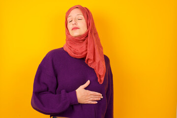 Satisfied smiling Young caucasian Muslim woman wearing hijab, keeps hands on belly, being in good mood after eating delicious supper, demonstrates she is full. Pleasant feeling in stomach.