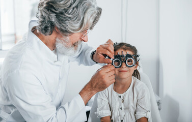 Senior male ophthalmologist with grey hair and beard in white coat is indoors in clinic testing sight of little girl