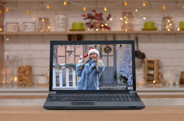 Safe Online greetings for the Christmas holidays ! Laptop on wooden table on kitchen background .Happy girl in a Santa hat waving hello on the background of the Christmas house on the monitor screen!
