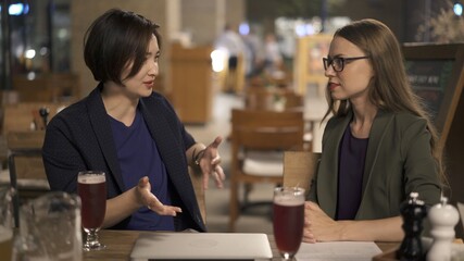 Two women friends sitting at the bar, blond woman in eyeglasses sitting on background of empty chairs. Two women discussing in an open space bar