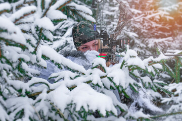 A young beautiful hunter woman in winter camouflage with hunting bow in her hand. All around are snowy trees.
