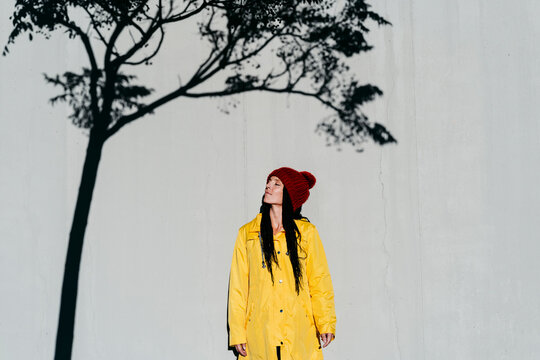 Woman wearing raincoat standing with with eyes closed under tree shadow against wall