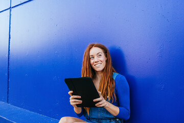Smiling redhead woman using digital tablet while sitting against blue wall