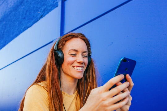 Redhead woman wearing headphones using mobile phone while sitting against blue wall