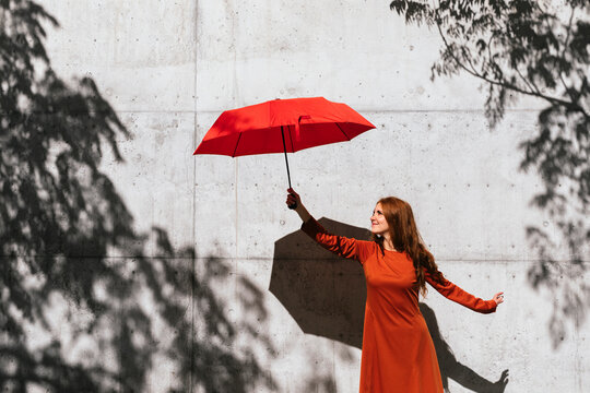 Smiling redhead woman holding umbrella while standing against tree shadow wall