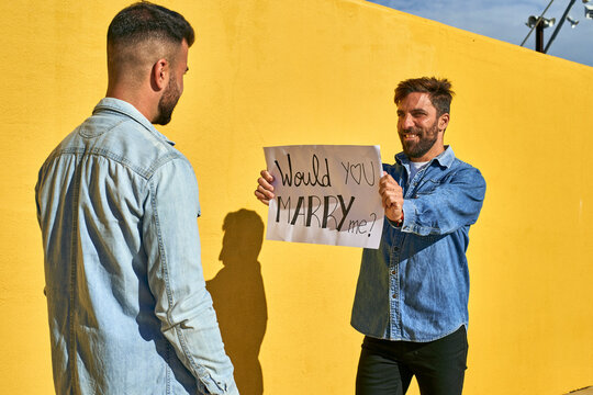 Man holding paper with written text Would You Marry Me on it while standing by yellow wall