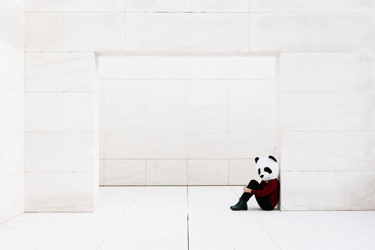 Woman wearing panda mask sitting at doorway against white wall