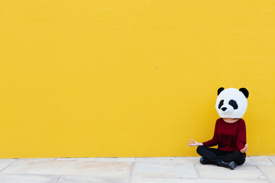 Woman wearing panda mask while sitting in lotus position against yellow wall