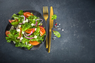 Bowl of healthy vegetable salad with various leaf salad, tomatoes, feta cheese, onions, olive oil, dark grey concrete background copy space