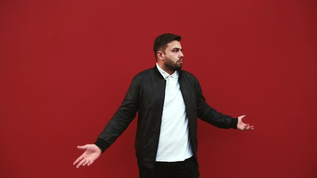 Confused bearded man in casual clothes stands against the background of a red wall and looks to the side with a puzzled face and spreads his arms.