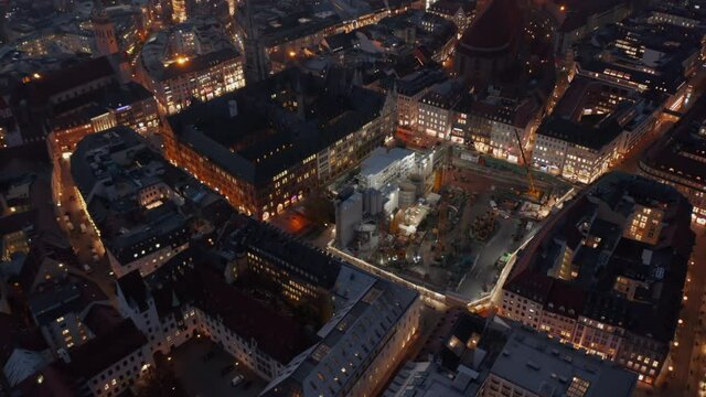 Huge Construction Building Site in Urban environment City Center of Munich, Germany at Night with lighting equipment and big machinery, Aerial dolly out tilt up