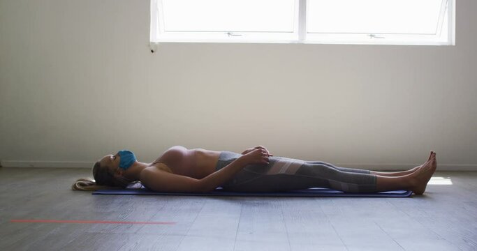 Fit caucasian woman wearing face mask performing yoga while lying in yoga studio