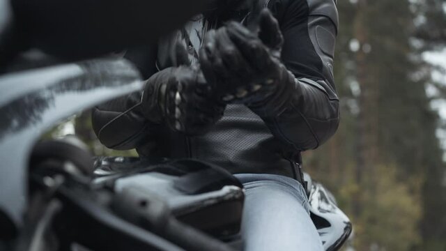 Unrecognizable motorcyclist putting on leather biker gloves sitting on motorbike. Young man preparing for speed racing outdoors on cloudy overcast day. Biking and masculinity concept.