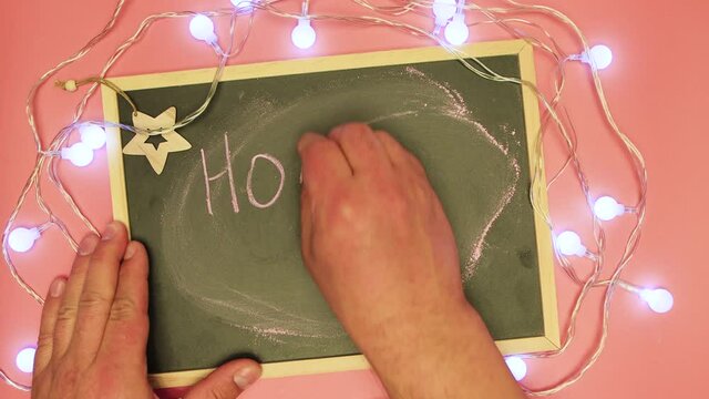 A man's hand writes Holly night on a black tablet. There is a garland and Christmas white lights are blinking. Dad makes a Happy New Year greeting manuscript. Flat laid holiday items.