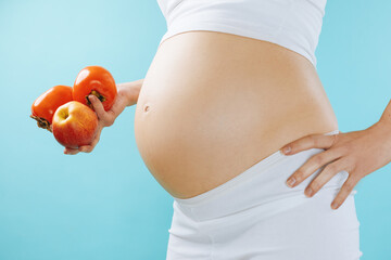 Pregnant woman with nude belly is holding fruits apple and persimmon isolated on blue background. 