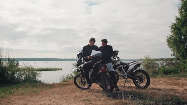 Tracking slowmo of young male motorcyclists resting by lake on gloomy day and looking at map