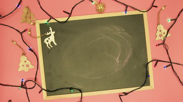 A man's hand writes Xo Xo Xo with chalk on a black board. There are wooden toys and Christmas colored lights are flashing. Dad makes a Happy New Year greeting manuscript. Flat laid holiday items.