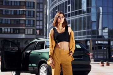Naklejka premium Modern business building at background. Young fashionable woman in burgundy colored coat at daytime with her car