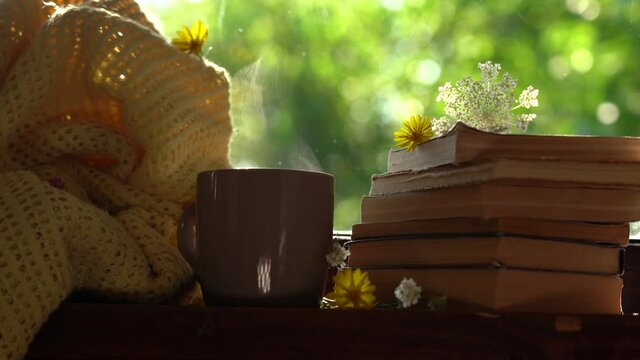 Steaming mug with hot tea or coffee isolated on green sunny blurry natural bokeh background. Old paper books stacked together on window sill. Cozy yellow knitted blanket.