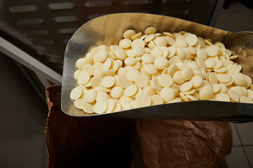 Granules of white chocolate raw materials in a metal ladle on the background of a small-scale wholesale line for the production sweets and confectionery