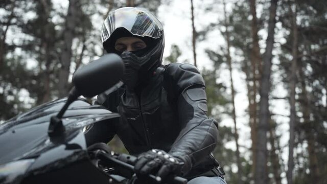 Young male biker in leather jacket and helmet sitting on black motorcycle and looking at camera. Portrait of confident handsome Middle Eastern man posing outdoors. Racing and hobby concept.