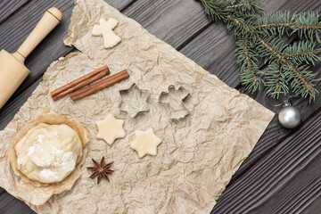 Fototapeta na wymiar Dough, molds, rolling pin and cinnamon sticks on paper. Spruce twigs on dark wooden background