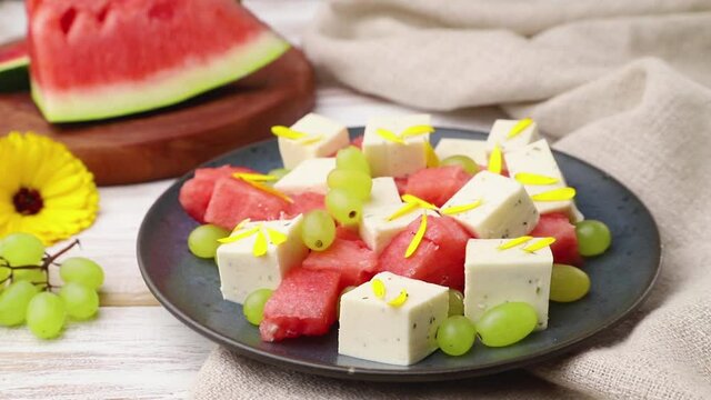 Vegetarian salad with watermelon, feta cheese, and grapes decorating with flower petals on blue ceramic plate on white wooden background and linen textile. Side view, close up, selective focus.
