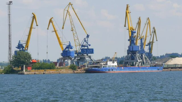 A row of construction or cargo cranes on the banks of a river or pond against the background of a barge standing nearby