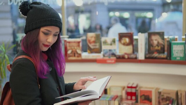 Portrait of stunning asian woman with bright red lips and purple hair reading a book in a bookstore. Millenials lifestyle. teenage student shopping. Concept of hipster style and trends.