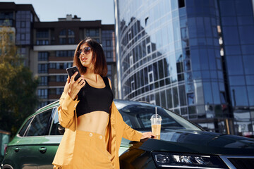 With smartphone. Young fashionable woman in burgundy colored coat at daytime with her car