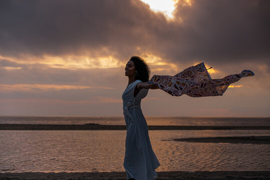 Happy Ethnic Young Woman Enjoying Life On Ocean Beach At Twilight In Windy Weather