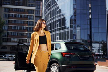 Fototapeta na wymiar Walks forward from automobile. Young fashionable woman in burgundy colored coat at daytime with her car