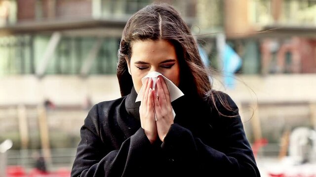 Eine junge Frau mit dunklen Haare schn&auml;uzt sich, putz sich ihre Nase mit Hilfe  von einem Taschentuch drau&szlig;en in einer Stadt.