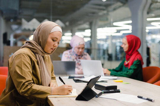 Pensive Islamic businesswoman writing in notebook in modern workplace