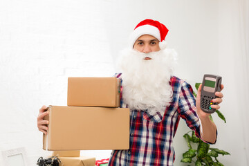 Fototapeta premium Positive smiling male courier in uniform and santa claus hat holding payment terminal and paper shopping bags, easy express order and delivery. Indoor studio