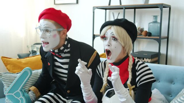 Man and woman mimes are watching sports on TV at home holding football and foam hand cheering then celebrating victory hugging kissing having fun