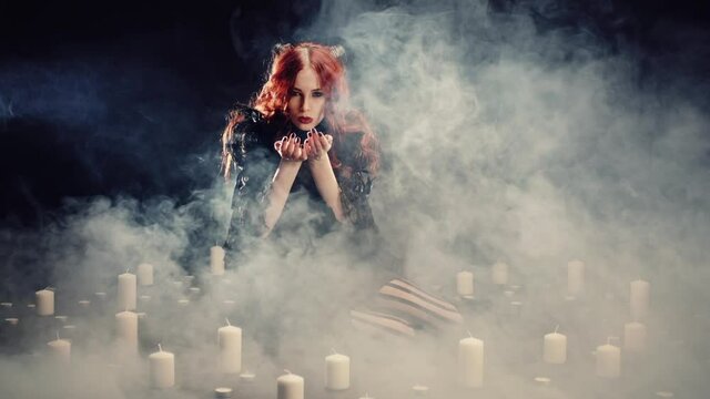 Young red-haired girl with devil horns and in black suit in dark room with smoke sitting on floor and causing evil spirits. Woman holding ancient magic occult ritual. Concept of mystic, supernatural.