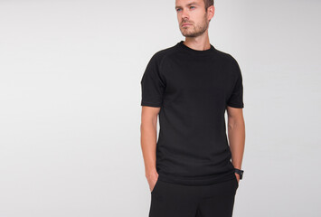 young stylish guy in black t-shirt and black pants is standing straight and looking away on the white wall background. medical concept. free space on left side, mockup