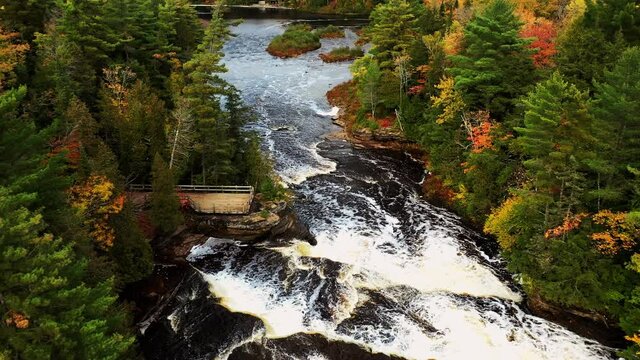 Aerial flying down the Tahquamenon River toward the lower waterfall cascades and observation deck situated between a forest of evergreen and autumn foliage covered trees.