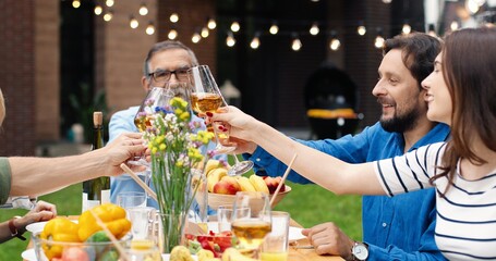 Cheerful happy Caucasian family sitting at table outdoors, having dinner and cheering with drinks while toasting. Joyful parents with children spending weekend together and celebrating.